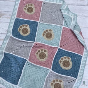 May include: A handmade crochet blanket featuring a patchwork design with squares in shades of blue, pink, and gray. Each square has a paw print in the center. The blanket has a white and light blue border.