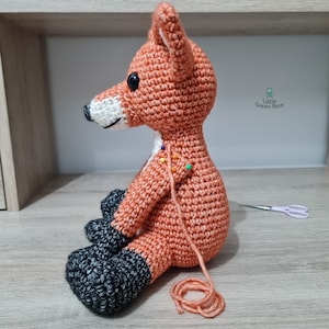 May include: A crocheted fox toy with orange fur, black feet, and white facial details. The toy is sitting on a wooden surface with a spool of orange yarn and a pair of scissors.
