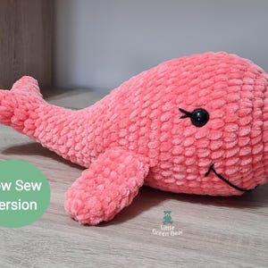 May include: A pink crocheted whale plush toy with black eyes and a stitched smile. The toy is lying on a wooden surface. The text "Low Sew Version" is visible in the image.