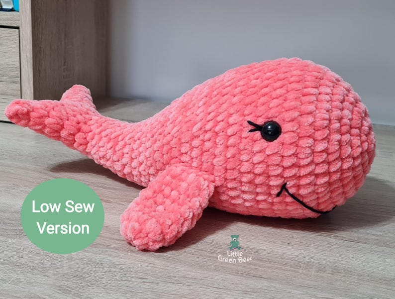 May include: A pink crocheted whale plush toy with black eyes and a stitched smile. The toy is lying on a wooden surface. The text "Low Sew Version" is visible in the image.