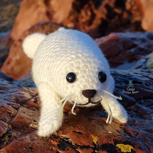 May include: A white crocheted seal plush toy with black plastic eyes, a dark brown stitched nose, and white thread whiskers. The toy features a small logo on the side with the text Little Green Bear and a small bear icon.