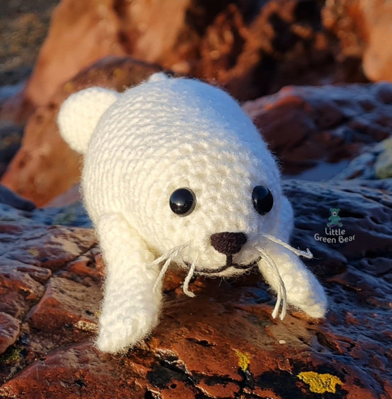 May include: A white crocheted seal plush toy with black plastic eyes, a dark brown stitched nose, and white thread whiskers. The toy features a small logo on the side with the text Little Green Bear and a small bear icon.