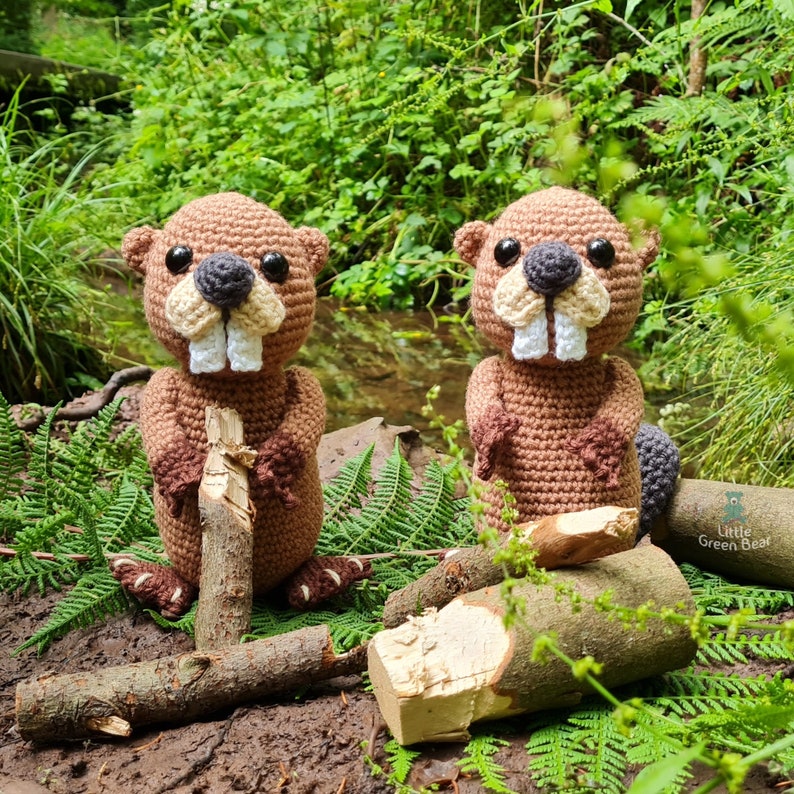 May include: Two brown crocheted beaver plushies with white teeth and black eyes are sitting on a bed of green ferns and brown branches. The beavers are holding sticks in their paws.