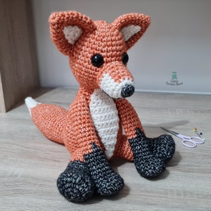 May include: A crocheted fox plush toy with orange and white fur, black eyes, and black feet. The fox is sitting on a wooden surface.