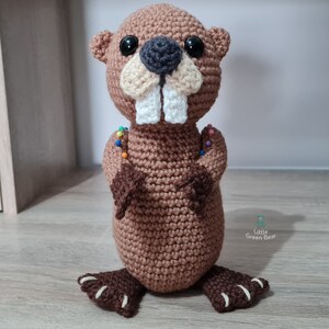 May include: A brown crocheted beaver pincushion with white teeth, black eyes, and a beige nose. The pincushion has rainbow coloured pins sticking out of it. The pincushion is standing on a wooden surface. The text "Little Green Bear" is visible in the lower right corner of the image.