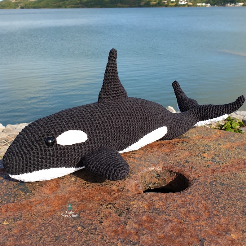 May include: A black and white crocheted orca plush toy featuring a prominent dorsal fin, a rounded tail, and a small logo with the text Little Green Bear printed on the side. The orca has a smooth, textured yarn surface and black bead eyes.