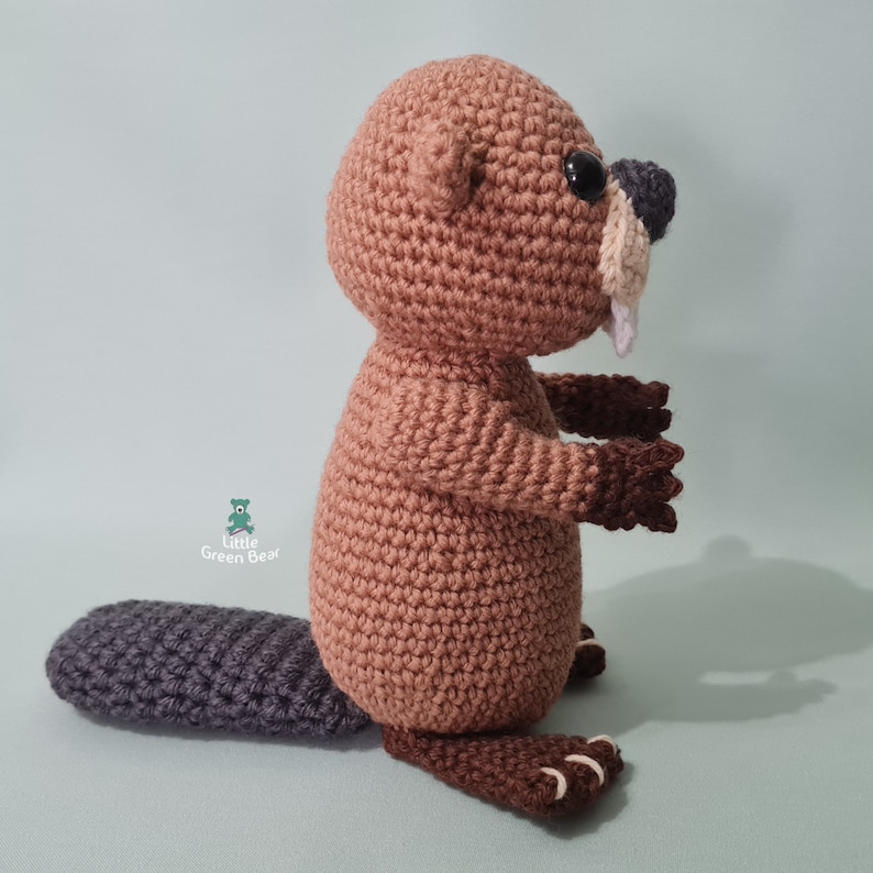 May include: A brown crocheted beaver with a grey tail, black eyes, and a white tooth. The beaver is standing on a light blue surface.
