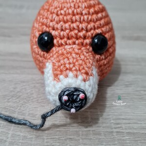 May include: Close-up of a crocheted fox head with black eyes and a black nose. The nose is being sewn with black yarn and has pink beads around it.