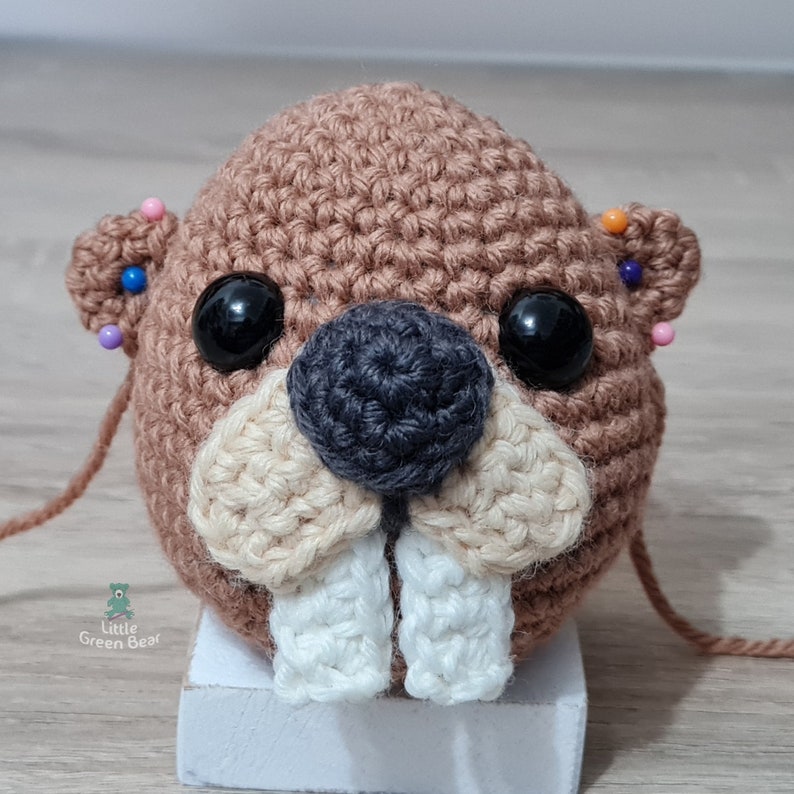 May include: A brown crocheted beaver with white teeth, black nose, and black eyes. The beaver is pinned with colourful pins.