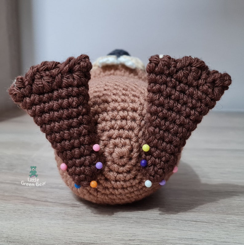 May include: A brown crocheted stuffed animal with brown ears, pinned together with colourful pins. The animal is lying on its back with its belly facing the camera.
