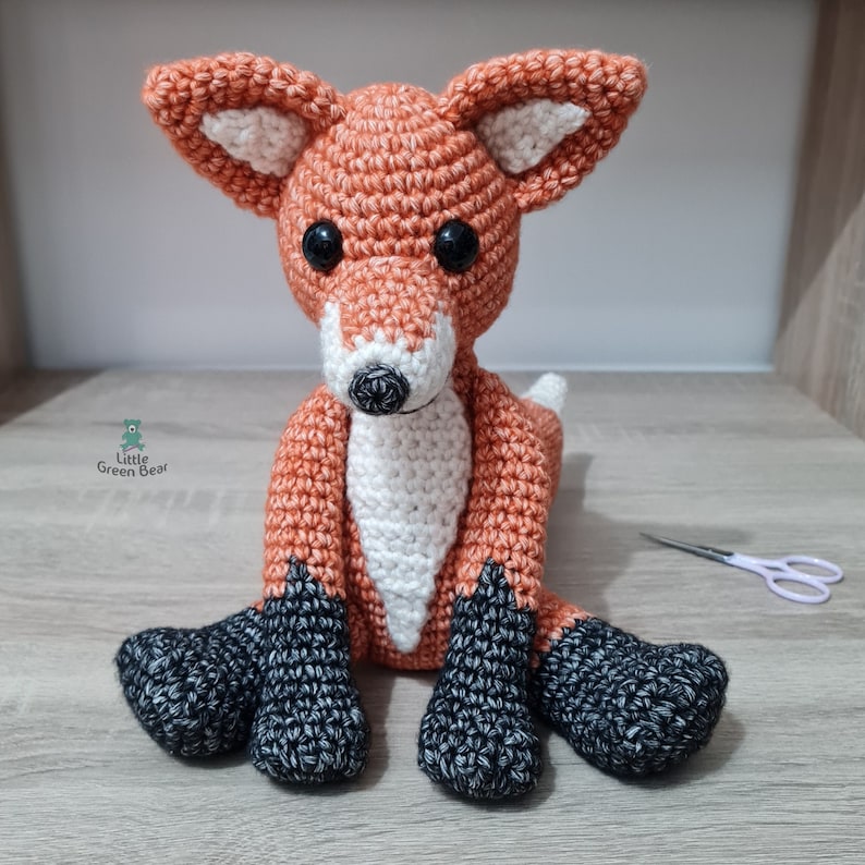 May include: A crocheted fox plush toy with orange fur, white chest, and black feet. The toy is sitting on a wooden surface.