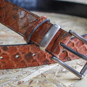 K&ouml;nnte beinhalten: Ein brauner G&uuml;rtel mit einem strukturierten, schuppenartigen Muster auf dem Riemen. Der G&uuml;rtel verf&uuml;gt &uuml;ber eine rechteckige Metallschnalle mit mattem Finish und eine passende Lederschlaufe. Die Kanten sind dunkel vern&auml;ht und die Innenseite ist schwarz.