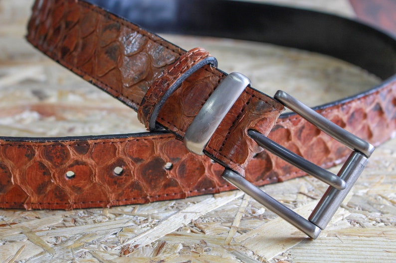 K&ouml;nnte beinhalten: Ein brauner G&uuml;rtel mit einem strukturierten, schuppenartigen Muster auf dem Riemen. Der G&uuml;rtel verf&uuml;gt &uuml;ber eine rechteckige Metallschnalle mit mattem Finish und eine passende Lederschlaufe. Die Kanten sind dunkel vern&auml;ht und die Innenseite ist schwarz.