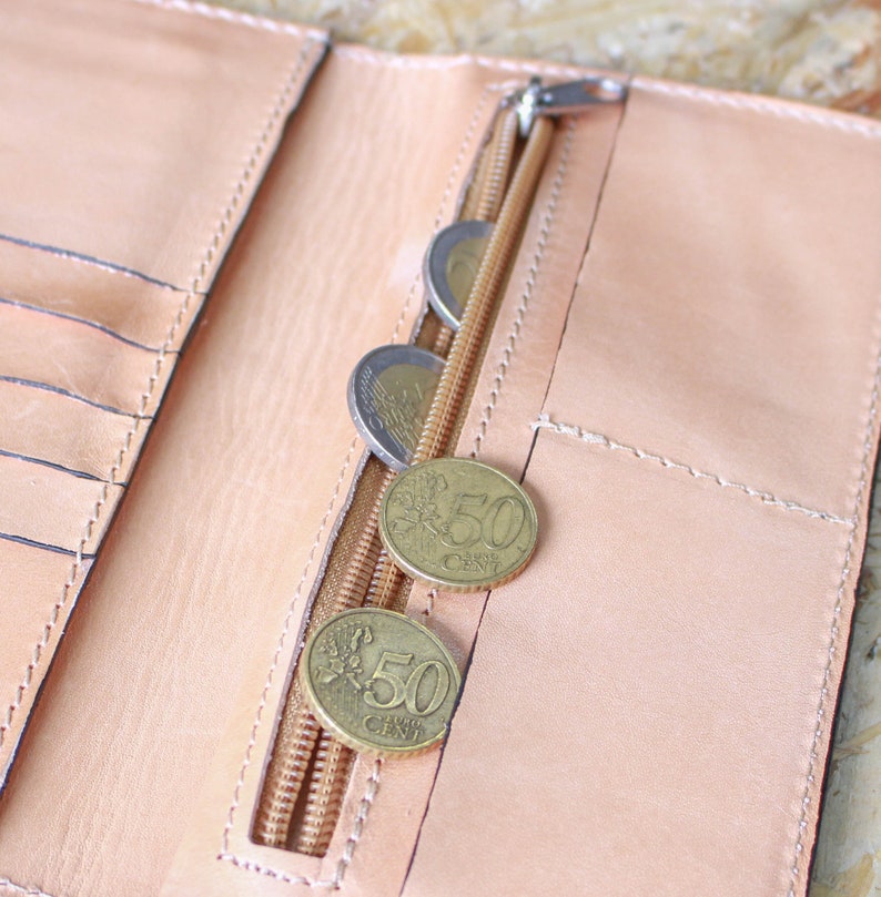 May include: A light brown leather wallet with a zippered pocket and several euro coins inside. The coins are 50 cent pieces.