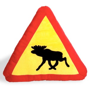 Puede incluir: Una almohada triangular amarilla con un borde rojo y una silueta negra de un alce. La almohada tiene forma de señal de advertencia.