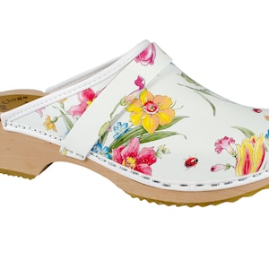 Pode incluir: Clog de couro branco com estampa floral e salto de madeira. O clog tem uma tira na parte superior e uma sola branca.