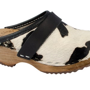 Puede incluir: Zuecos de piel de vaca en blanco y negro con una tira de cuero negro y un tacón de madera. Los zuecos tienen una costura blanca alrededor del borde.