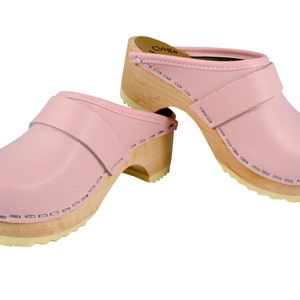 May include: A pair of pink leather clogs with a wooden heel and a strap across the top. The clogs have a stitched seam around the edge of the sole.