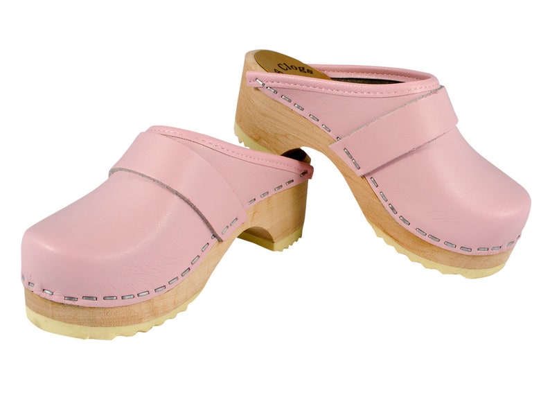May include: A pair of pink leather clogs with a wooden heel and a strap across the top. The clogs have a stitched seam around the edge of the sole.