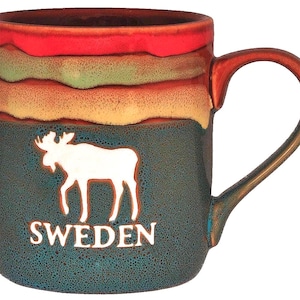 May include: A ceramic mug with a white moose silhouette and the word "SWEDEN" on a teal background. The top of the mug features bands of red, green, orange, and tan. The handle and rim are brown.
