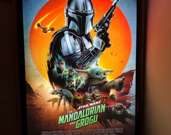 May include: A rectangular framed poster for The Mandalorian and Grogu featuring a central figure in a silver helmet holding a blaster, with Grogu below, set against a large orange sun. The text reads The Mandalorian and Grogu, Only in Theaters May 22.