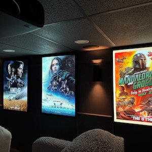 May include: Three rectangular, illuminated movie posters mounted on a dark wall. The left poster features Andor, the center poster displays Rogue One, and the right poster shows The Mandalorian and Grogu with text including Only in theatres MAY 22.