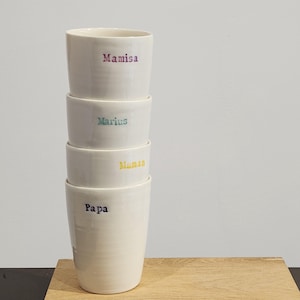 Peut inclure: Quatre tasses en céramique blanc cassé empilées, avec des noms en lettrage coloré : "Mamisa", "Marius", "Maman" et "Papa". Les tasses sont cylindriques et ont une finition lisse et brillante. Elles sont disposées sur une surface en bois brun clair.