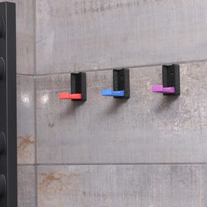 Op de afbeelding: Drie aan de muur gemonteerde haken met een zwarte basis en gekleurde accenten. De haken zijn rood, blauw en paars. Ze zijn gemonteerd op een grijze betegelde muur, in de buurt van een zwarte handdoekrek.