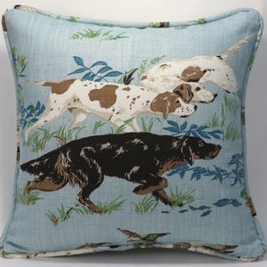 May include: Square cushion with a light blue background featuring a repeating pattern of three dogs in a field. The dogs are brown and white, and black and brown. The cushion has a light blue border.