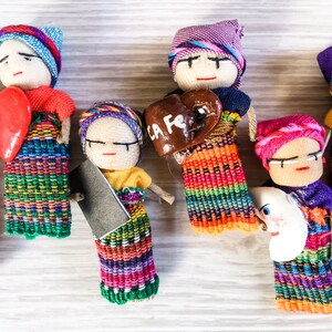 Puede incluir: Una colecci&oacute;n de mu&ntilde;ecas artesanales coloridas con ropa bordada y accesorios &uacute;nicos. Las mu&ntilde;ecas sostienen un coraz&oacute;n rojo, un coraz&oacute;n marr&oacute;n con el texto "La Fe" y una luna creciente blanca.