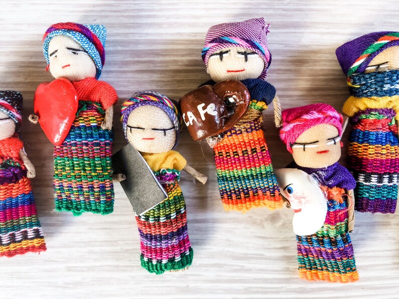 Puede incluir: Una colecci&oacute;n de mu&ntilde;ecas artesanales coloridas con ropa bordada y accesorios &uacute;nicos. Las mu&ntilde;ecas sostienen un coraz&oacute;n rojo, un coraz&oacute;n marr&oacute;n con el texto "La Fe" y una luna creciente blanca.