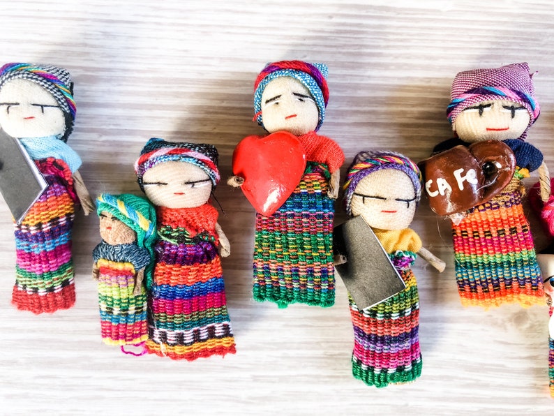 Puede incluir: Un conjunto de seis mu&ntilde;ecas artesanales coloridas con ropa y rostros bordados. Las mu&ntilde;ecas visten ropa tradicional guatemalteca y sostienen varios objetos, entre ellos un coraz&oacute;n, una taza y un libro.