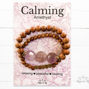 Puede incluir: Un conjunto de dos pulseras con cuentas de amatista y cuentas de madera. Las pulseras están apiladas una encima de la otra y tienen un acento dorado. La tarjeta dice "Calming Amethyst" y "relaxing peaceful healing".