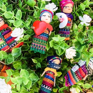 Puede incluir: Una colecci&oacute;n de mu&ntilde;ecas tejidas de colores con varios accesorios, incluyendo un coraz&oacute;n, una luna y una mu&ntilde;eca peque&ntilde;a. Las mu&ntilde;ecas est&aacute;n dispuestas sobre un lecho de hojas verdes y flores blancas.
