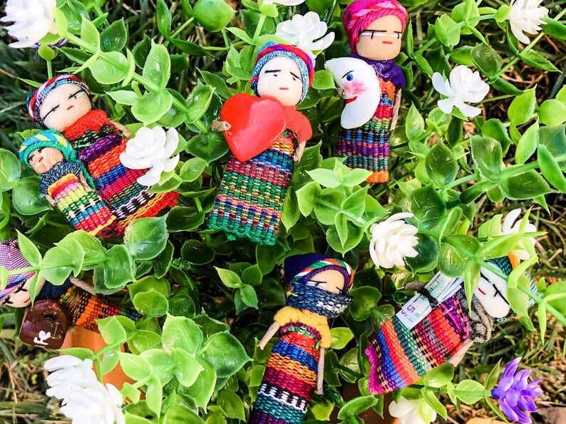 Puede incluir: Una colecci&oacute;n de mu&ntilde;ecas tejidas de colores con varios accesorios, incluyendo un coraz&oacute;n, una luna y una mu&ntilde;eca peque&ntilde;a. Las mu&ntilde;ecas est&aacute;n dispuestas sobre un lecho de hojas verdes y flores blancas.