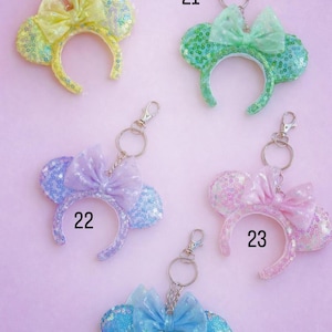 May include: A selection of keychains with sequined mouse ear headbands in yellow, green, purple, pink, and blue. Each headband has a matching bow and a silver clasp for attaching to keys or bags. Numbers 20, 21, 22, 23, and 25 are visible.