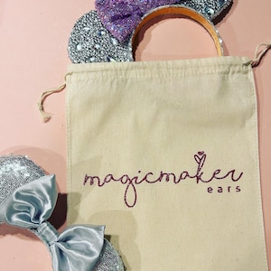 May include: A pair of silver glitter mouse ears with a purple bow and a silver satin bow, in a white drawstring bag with the text "magicmaker ears" in pink glitter.