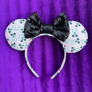 May include: A Halloween-themed headband with mouse ears. The ears are light pink with a repeating pattern of a ghost and Mickey Mouse. A large black sequined bow sits between the ears. The headband is white with the same ghost and Mickey Mouse pattern.