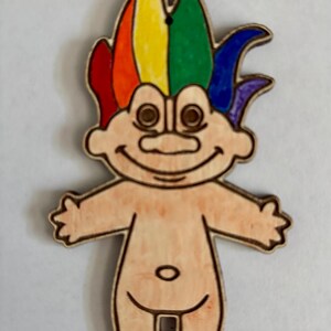May include: Wooden troll figurine with rainbow hair. The troll has a smiling face, outstretched arms, and a small belly button. The hair is colored red, orange, yellow, green, blue, and purple. The figurine is made of light-colored wood.