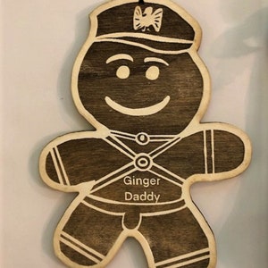 May include: Wooden gingerbread man ornament with a smiling face, wearing a hat with a decorative emblem. The ornament has the words "Ginger Daddy" etched on the front. A festive holiday decoration.