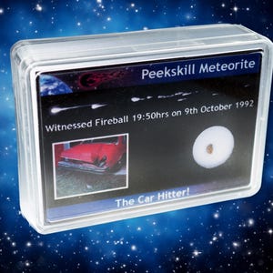 May include: A clear plastic case containing a Peekskill Meteorite. The display card features text about a witnessed fireball and a photo of a red car. The text reads "The Car Hitter!"