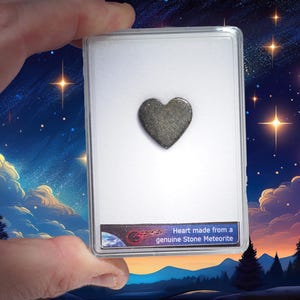 Pode incluir: Um meteorito de pedra em forma de coração em uma vitrine de plástico transparente sobre um fundo branco. A vitrine tem uma etiqueta que diz "Coração feito de um meteorito de pedra genuíno".