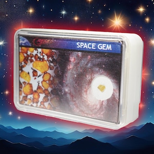 Pode incluir: Uma caixa de plástico branca com uma janela de plástico transparente que mostra uma pequena rocha branca com um cristal amarelo dentro. A caixa tem as palavras "SPACE GEM" impressas na frente.