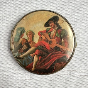 May include: A round, vintage compact with a painted scene of a man playing a guitar for two women. The man wears a black hat and red and green clothing. The compact has a gold-colored metal frame.