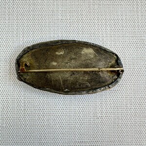 May include: An antique oval brooch with a tarnished silver-tone metal frame. The pin has a gold-tone clasp and pin. The brooch is set against a textured, off-white background.