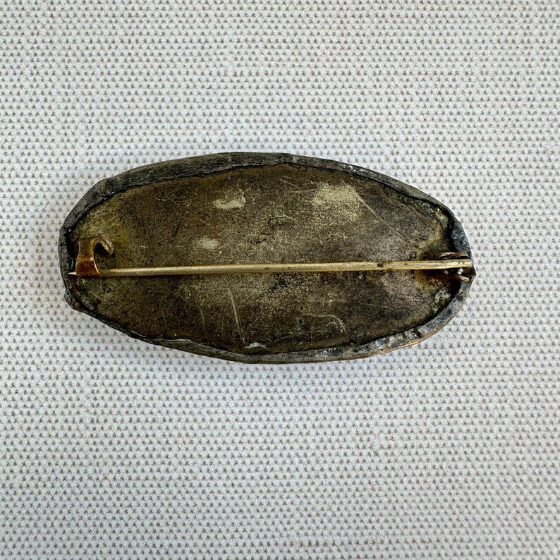 May include: An antique oval brooch with a tarnished silver-tone metal frame. The pin has a gold-tone clasp and pin. The brooch is set against a textured, off-white background.