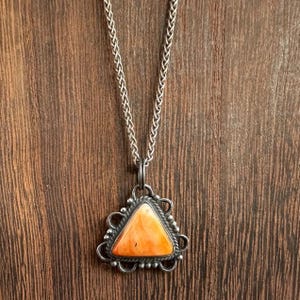 May include: A silver necklace with a triangular pendant. The pendant features an orange stone set in a decorative silver frame. The chain is a woven style, and the background is a wooden surface.
