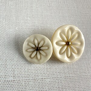 May include: Two round off-white buttons of different sizes, each featuring a molded floral design with eight petals radiating from a small, dark metallic center. The buttons have a smooth, glossy finish and a slightly raised, rounded edge profile.