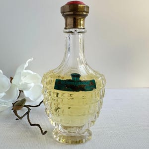 May include: A vintage clear glass perfume bottle with a gold-coloured cap and a red top. The bottle has a unique tiered design and contains a light yellow liquid. A green and blue label reads "Eau de Cologne Double".