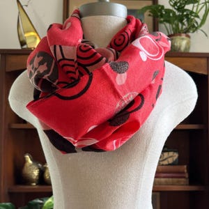 Könnte beinhalten: Ein roter Loop-Schal mit einem sich wiederholenden Muster aus schwarzen, weißen und braunen abstrakten Formen. Der Schal wird auf einer Schaufensterpuppe präsentiert und scheint aus einem weichen Stoff zu sein.